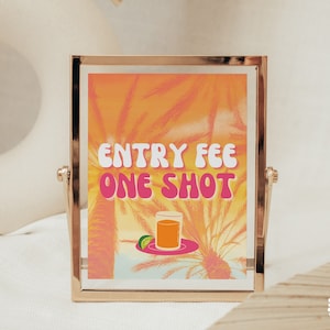 May include: A gold-framed sign with a tropical background and the text "ENTRY FEE ONE SHOT" in pink letters. A glass of orange juice with a lime wedge sits on a plate in front of the text.