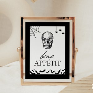 Puede incluir: Un letrero con marco dorado, fondo negro y texto blanco que dice "Bone Appetit". El letrero presenta una ilustración en blanco y negro de una calavera con una telaraña y murciélagos.