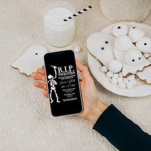 May include: A black smartphone with a white skeleton holding a drink and the text "RIP TWENTIES" on the screen. The phone is being held by a hand wearing a black sleeve. The background is a white fuzzy surface with white ghost-shaped cookies and white marshmallows.