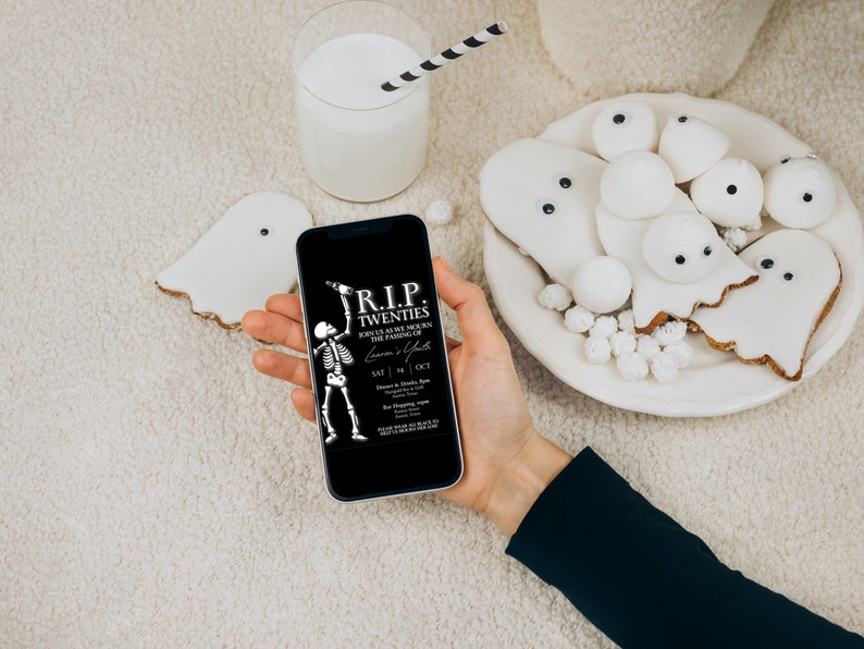 May include: A black smartphone with a white skeleton holding a drink and the text "RIP TWENTIES" on the screen. The phone is being held by a hand wearing a black sleeve. The background is a white fuzzy surface with white ghost-shaped cookies and white marshmallows.