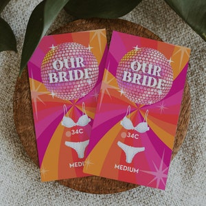 May include: Two pink and orange cards with a disco ball design. The cards say "Our Bride" and feature a white bra and underwear with the size "34C" and "Medium" printed on the cards.