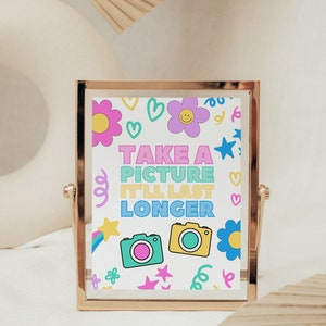 Peut inclure: Une image encadrée en or avec un fond blanc et des illustrations colorées de fleurs, d'étoiles et de confettis. Le texte "TAKE A PICTURE IT'LL LAST LONGER" est écrit en couleurs arc-en-ciel. Deux caméras de dessin animé se trouvent en bas de l'image.
