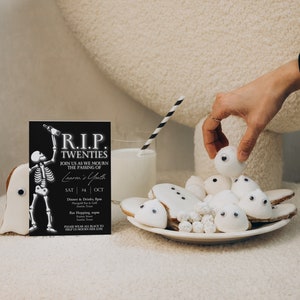 May include: A black and white invitation card with the text "R.I.P. TWENTIES" and a skeleton holding a bottle. The invitation is for a party celebrating the end of someone's twenties. The card is sitting on a table with a plate of white ghost-shaped cookies with googly eyes.
