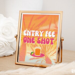 Puede incluir: Un cartel con marco dorado con un fondo de puesta de sol y palmeras. El cartel dice "Entry Fee One Shot" en letras rosas y blancas. Un vaso de l&iacute;quido naranja se encuentra en un plato con una rodaja de lima.