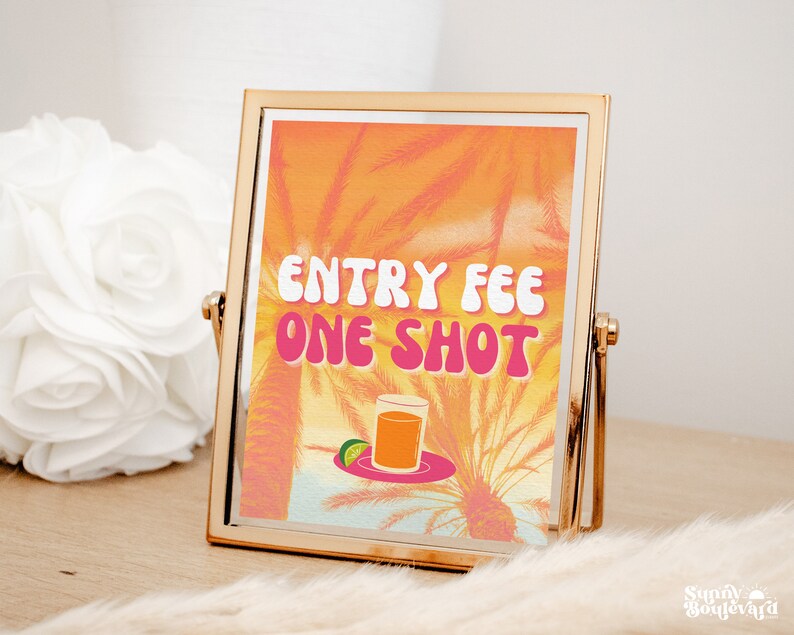 Puede incluir: Un cartel con marco dorado con un fondo de puesta de sol y palmeras. El cartel dice "Entry Fee One Shot" en letras rosas y blancas. Un vaso de l&iacute;quido naranja se encuentra en un plato con una rodaja de lima.
