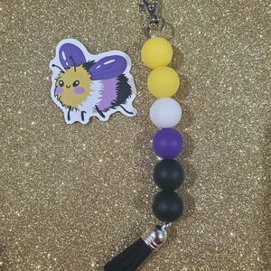 May include: A keychain with a non-binary pride flag design. The keychain features yellow, white, purple, and black beads, a silver-colored clasp, and a black tassel. A sticker of a cartoon bee with purple wings and a yellow body is also present.