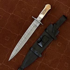 K&ouml;nnte beinhalten: Ein silbernes Dolch mit einer gemusterten Klinge und einem wei&szlig;en Hirschhorn-Griff. Der Dolch befindet sich in einer schwarzen Lederscheide mit einem Riemen.
