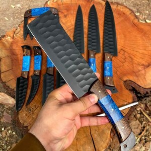 May include: A set of black kitchen knives with blue and brown handles. The knives have a textured blade and are arranged on a wooden surface.