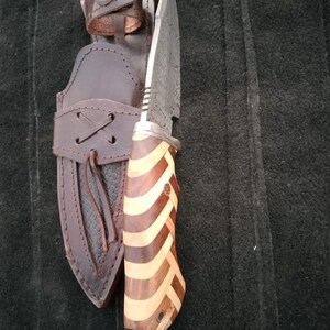 May include: A hunting knife with a patterned steel blade and a wooden handle with a striped pattern. The knife is in a brown leather sheath.