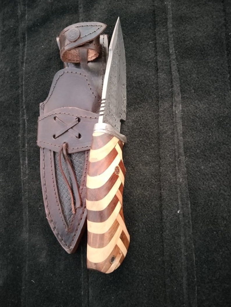 May include: A hunting knife with a patterned steel blade and a wooden handle with a striped pattern. The knife is in a brown leather sheath.