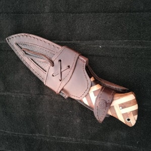 May include: A brown leather sheath with a wooden-handled knife with a geometric pattern. The sheath has a strap with a decorative knot.