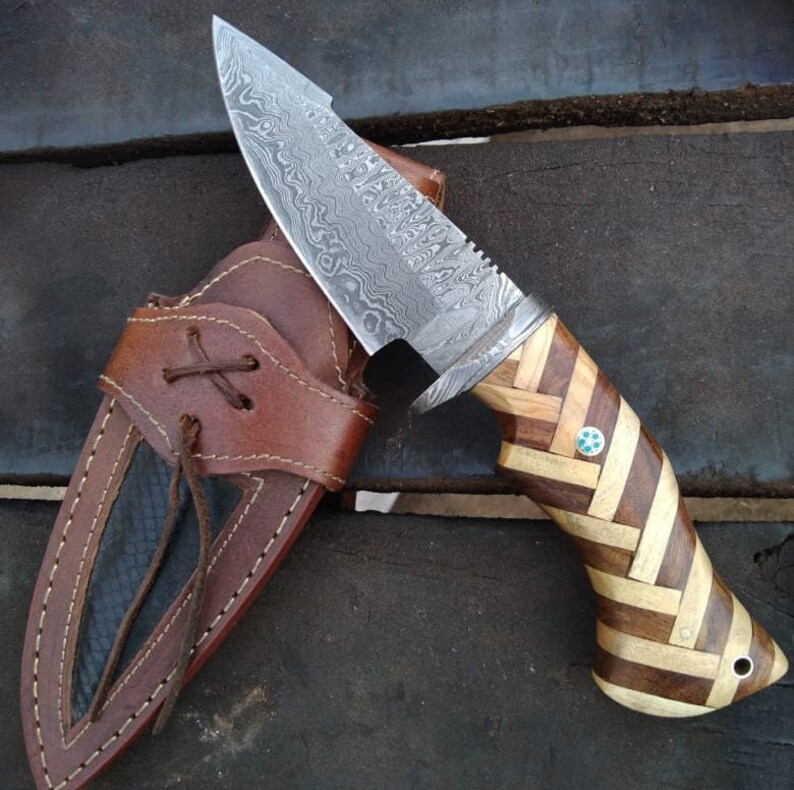 May include: A hunting knife with a Damascus steel blade and a wooden handle. The handle is made of multiple pieces of wood that are arranged in a herringbone pattern. The knife is in a brown leather sheath.