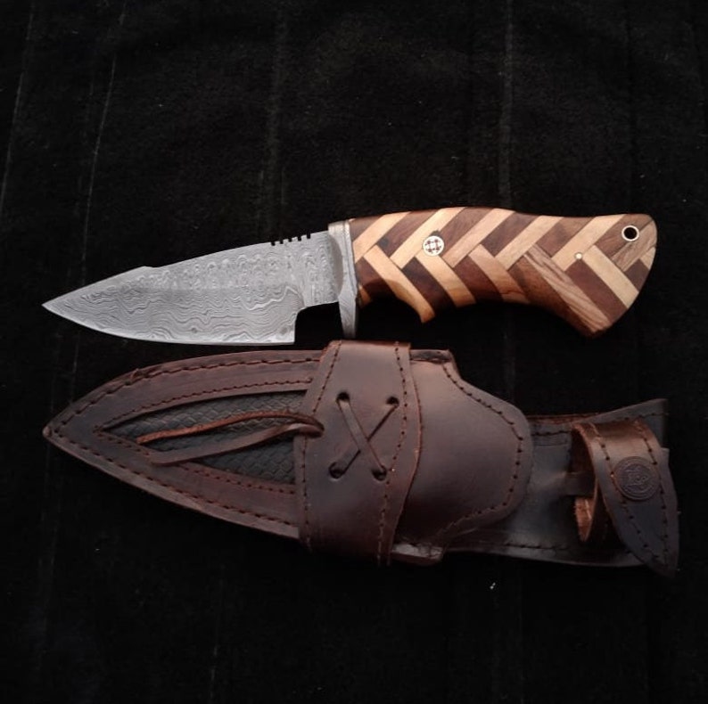 May include: A brown leather sheath with a decorative stitching pattern holds a hunting knife with a patterned wooden handle and a Damascus steel blade.