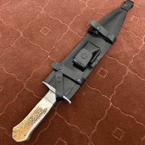 K&ouml;nnte beinhalten: Eine schwarze Lederscheide mit einem braunen Griffmesser. Das Messer hat einen wei&szlig;en Griff mit einem strukturierten Muster. Die Scheide hat einen Riemen und einen Schnallenverschluss.