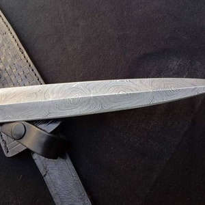 K&ouml;nnte beinhalten: Nahaufnahme eines silbernen Dolches mit einer gemusterten Klinge. Der Dolch befindet sich in einer schwarzen Lederscheide.