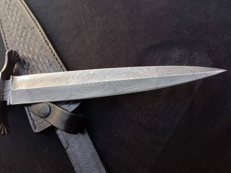 K&ouml;nnte beinhalten: Nahaufnahme eines silbernen Dolches mit einer gemusterten Klinge. Der Dolch befindet sich in einer schwarzen Lederscheide.