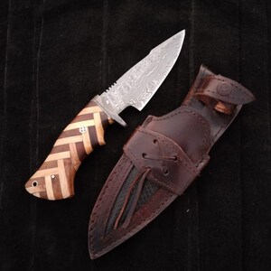 May include: A brown leather sheath with a black leather lining and a brown leather strap with a metal buckle. A knife with a wooden handle and a silver blade is in the sheath.