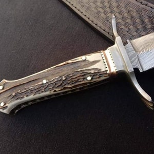 K&ouml;nnte beinhalten: Ein Jagdmesser mit einem braunen und wei&szlig;en Hirschhorn-Griff und einer silbernen Klinge mit einem dekorativen Muster. Das Messer liegt auf einer braunen Lederscheide.