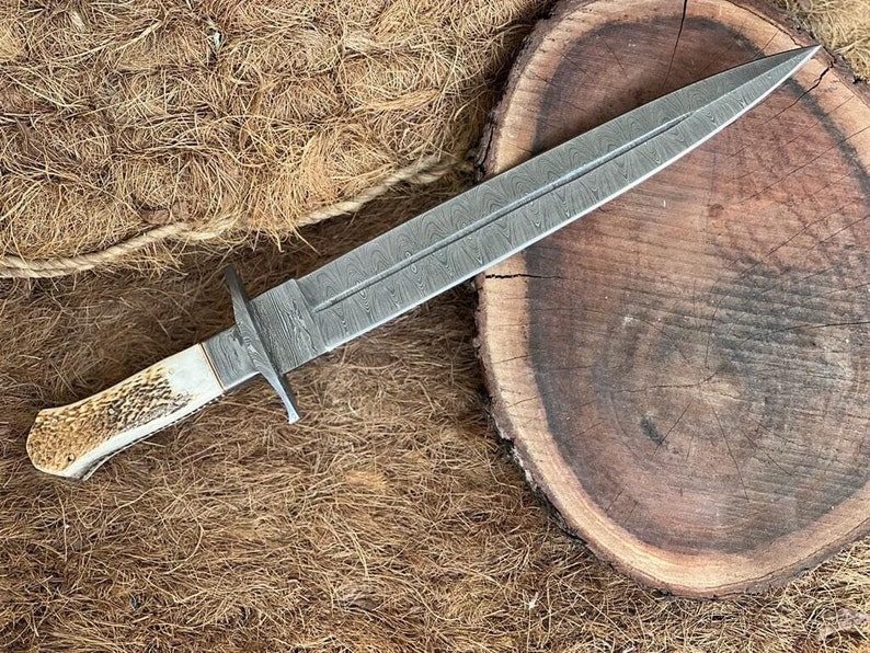 K&ouml;nnte beinhalten: Ein handgefertigtes Dolch mit einer Damaststahlklinge und einem Knochengriff. Die Klinge hat ein charakteristisches Muster und ist scharf. Der Griff ist aus Knochen und hat eine nat&uuml;rliche Oberfl&auml;che.