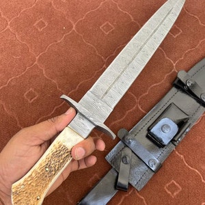 K&ouml;nnte beinhalten: Ein Damaststahl-Dolch mit einem wei&szlig;en Knochengriff und einer schwarzen Lederscheide. Die Klinge hat ein charakteristisches wellenf&ouml;rmiges Muster.
