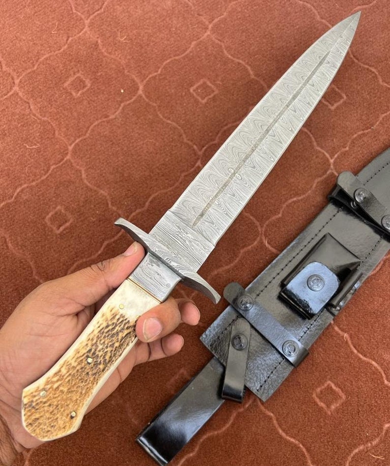 K&ouml;nnte beinhalten: Ein Damaststahl-Dolch mit einem wei&szlig;en Knochengriff und einer schwarzen Lederscheide. Die Klinge hat ein charakteristisches wellenf&ouml;rmiges Muster.