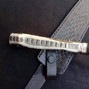 K&ouml;nnte beinhalten: Nahaufnahme eines Messergriffs mit einem dekorativen Muster aus Metall und Knochen. Der Griff ist an einer schwarzen Lederscheide mit einem gewebten Muster befestigt.