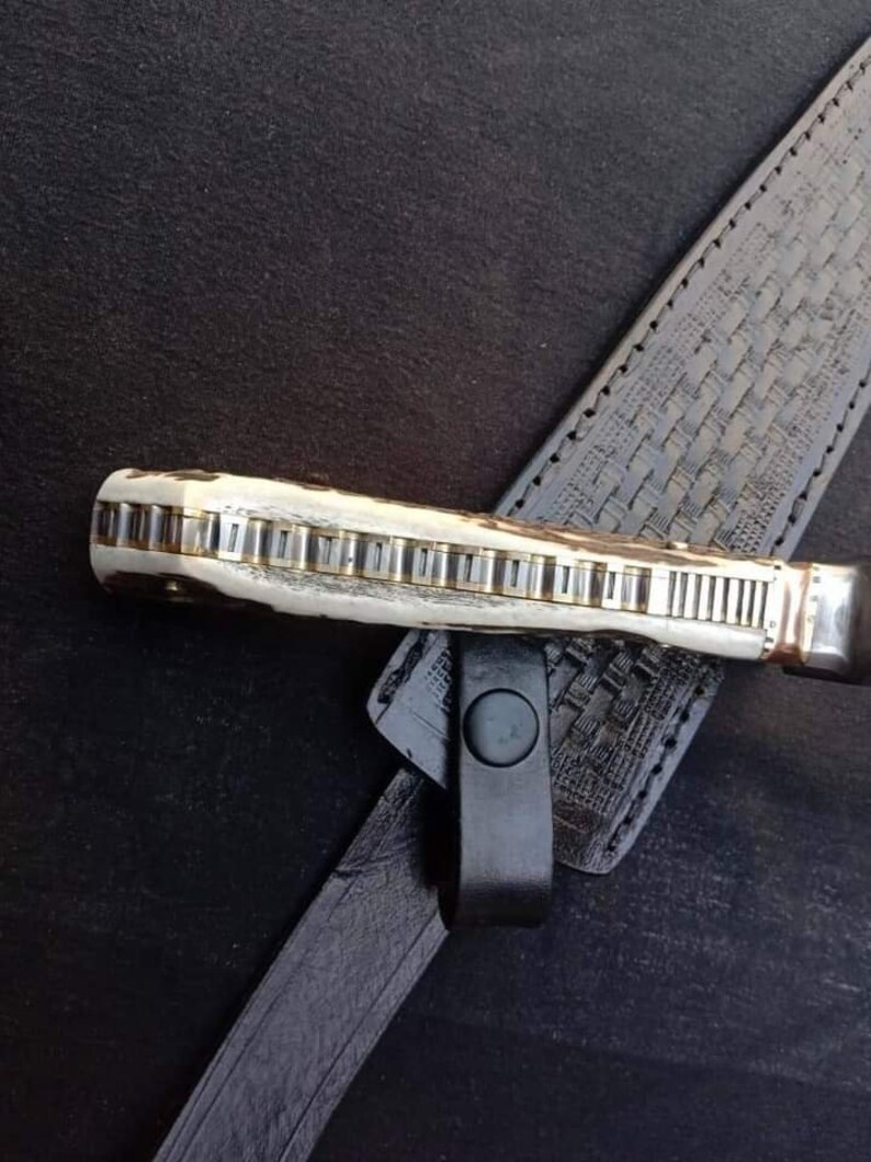 K&ouml;nnte beinhalten: Nahaufnahme eines Messergriffs mit einem dekorativen Muster aus Metall und Knochen. Der Griff ist an einer schwarzen Lederscheide mit einem gewebten Muster befestigt.