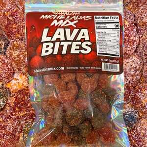 Può includere: Un sacchetto trasparente di caramelle "Lava Bites". La borsa ha un'etichetta rossa e nera con il testo "Shakalona Micheladas Mix Lava Bites". La borsa contiene molte caramelle, ciascuna ricoperta da una polvere marrone-rossastra. Il peso netto è di 170g.