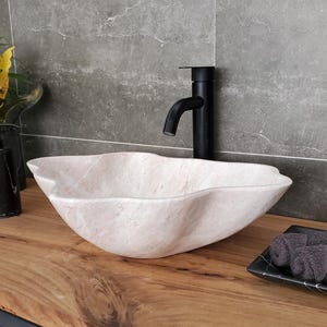 May include: A modern bathroom vanity with a light-colored stone vessel sink. A black faucet is mounted on the wall. A black soap dish with rolled gray towels sits on the wooden countertop. A black vase holds a plant with yellow and green leaves.