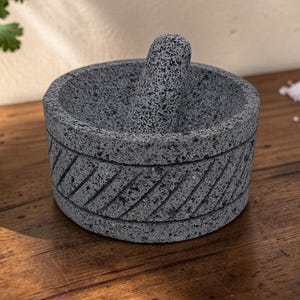 May include: A dark grey volcanic stone mortar and pestle set with a porous, textured surface. The cylindrical mortar features a decorative band of diagonal carved grooves around its exterior, while the pestle rests inside the bowl.