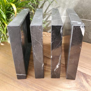 May include: Four dark gray marble bookends with white veining. The bookends are rectangular and stand upright on a wooden surface. The bookends are a decorative item for home or office.
