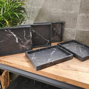 May include: A collection of rectangular serving trays with a black marble design and white veining. The trays are displayed on a wooden surface, showcasing their elegant and modern aesthetic. The trays vary in size.