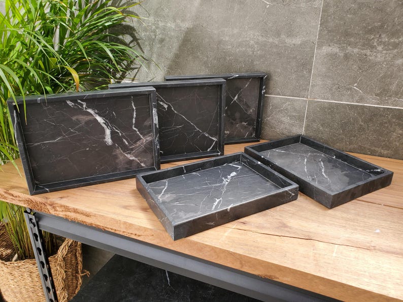 May include: A collection of rectangular serving trays with a black marble design and white veining. The trays are displayed on a wooden surface, showcasing their elegant and modern aesthetic. The trays vary in size.