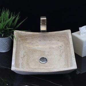 May include: A square, beige stone vessel sink with a curved edge and a drain in the center. The sink is on a black countertop with a plant in a gray pot and a white tissue box.