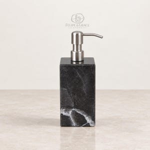 May include: A rectangular black marble soap dispenser with white veining and a brushed silver metal pump mechanism. The logo for Felipe & Grace Stone is printed in white above the dispenser. The base of the dispenser is square and solid.