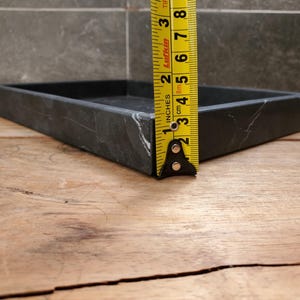 May include: A square, black marble tray with white veining. The tray is approximately 1 inch (2.5 cm) high. The tray is on a wooden surface, with a yellow tape measure next to it.