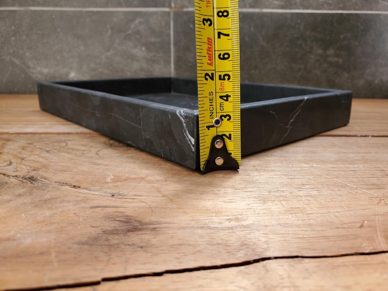 May include: A square, black marble tray with white veining. The tray is approximately 1 inch (2.5 cm) high. The tray is on a wooden surface, with a yellow tape measure next to it.