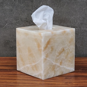 Puede incluir: Un dispensador de pañuelos cuadrado de piedra beige con vetas naturales. Los pañuelos blancos sobresalen de la abertura superior. La caja está sobre una superficie de madera contra un fondo gris oscuro. Un artículo decorativo para el hogar.