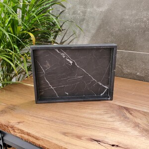 May include: A rectangular black marble tray with white veining. The tray has a raised edge and sits on a wooden surface. The tray is empty and ready to be used for serving or display.
