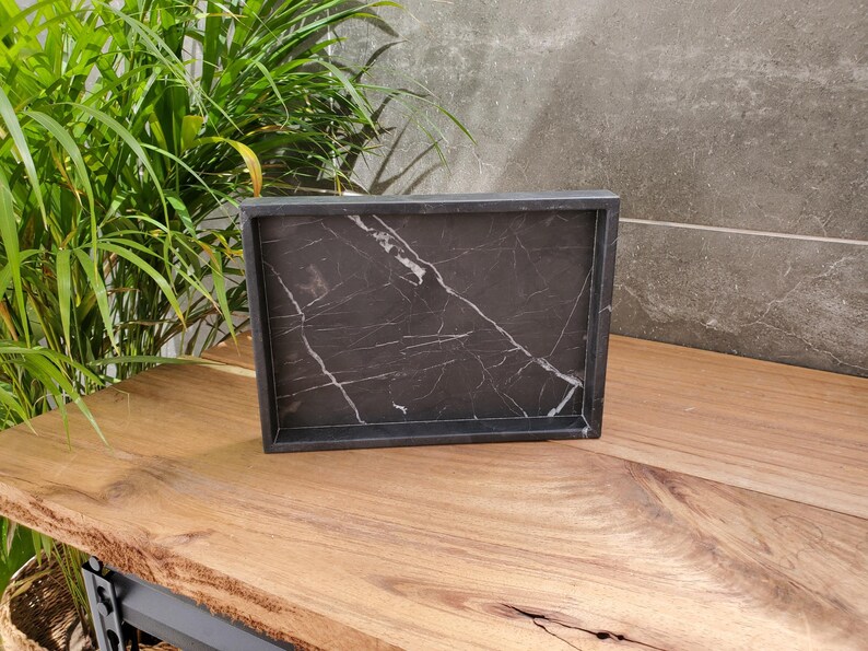 May include: A rectangular black marble tray with white veining. The tray has a raised edge and sits on a wooden surface. The tray is empty and ready to be used for serving or display.
