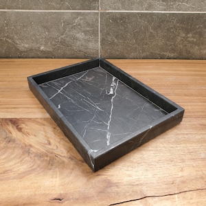 May include: A rectangular black marble tray with white veining. The tray has a raised edge and sits on a wooden surface. The background features a gray tiled wall. This tray is suitable for home decor.