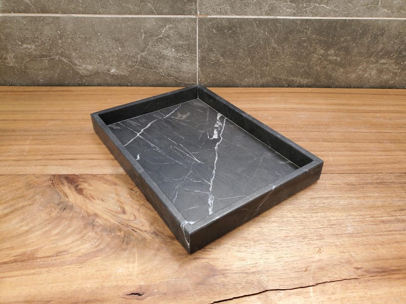 May include: A rectangular black marble tray with white veining. The tray has a raised edge and sits on a wooden surface. The background features a gray tiled wall. This tray is suitable for home decor.