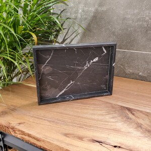 May include: A rectangular black marble tray with white veining. The tray has a raised border and sits on a wooden surface. The background includes a green plant and a gray wall. This tray is suitable for home decor.