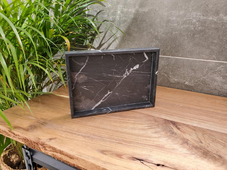 May include: A rectangular black marble tray with white veining. The tray has a raised border and sits on a wooden surface. The background includes a green plant and a gray wall. This tray is suitable for home decor.