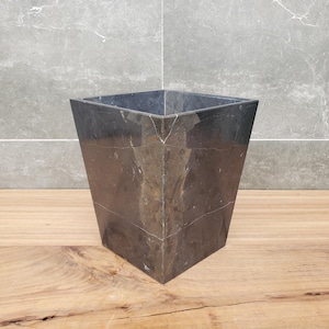 May include: A dark grey, square wastebasket made of stone. The sides are angled, and the corners are joined with visible seams. The stone has a marbled appearance with light-coloured veining. The wastebasket sits on a wooden surface.