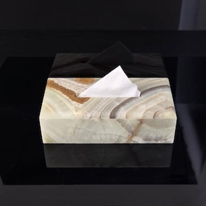 Puede incluir: Un dispensador de pañuelos rectangular hecho de piedra de ónix en capas. La caja presenta una mezcla de tonos blancos, marrones y verdes, con una parte superior de ónix negro. Un pañuelo blanco sobresale. Sobre una superficie negra reflectante.