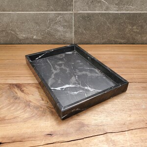 May include: A rectangular black marble tray with white veining. The tray has a raised edge and sits on a wooden surface. The marble has a polished finish, and the tray is empty.