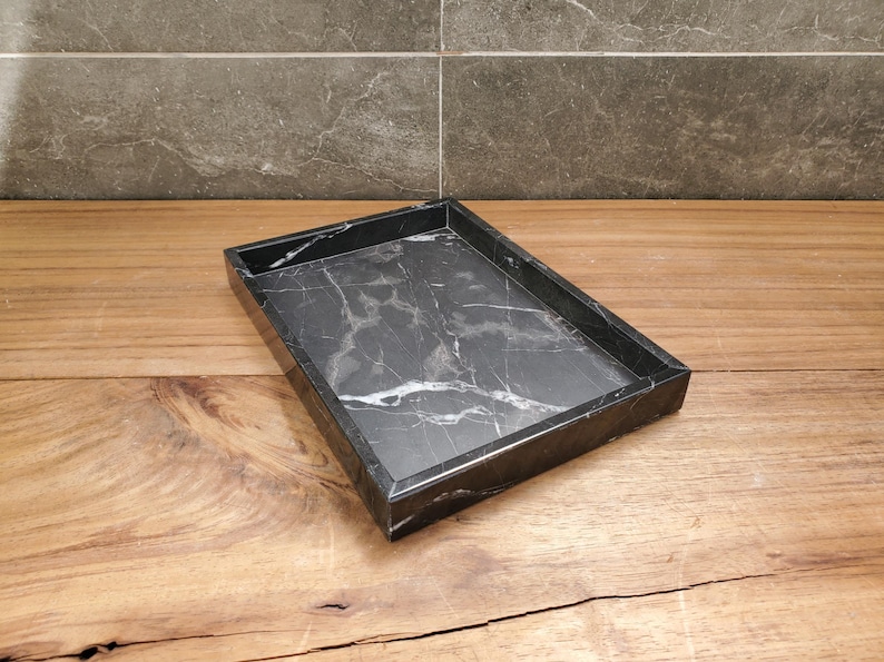 May include: A rectangular black marble tray with white veining. The tray has a raised edge and sits on a wooden surface. The marble has a polished finish, and the tray is empty.