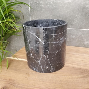 May include: A black marble container with white veining, cylindrical in shape. It sits on a wooden surface, with a green plant and a grey tiled wall in the background. The container has a smooth, polished finish.