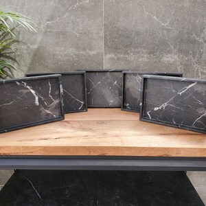 May include: Six rectangular serving trays with black marble-like surfaces and white veining are displayed on a wooden table. The trays have a dark frame and are arranged in a semi-circle. The background features a gray tiled wall and a green plant.